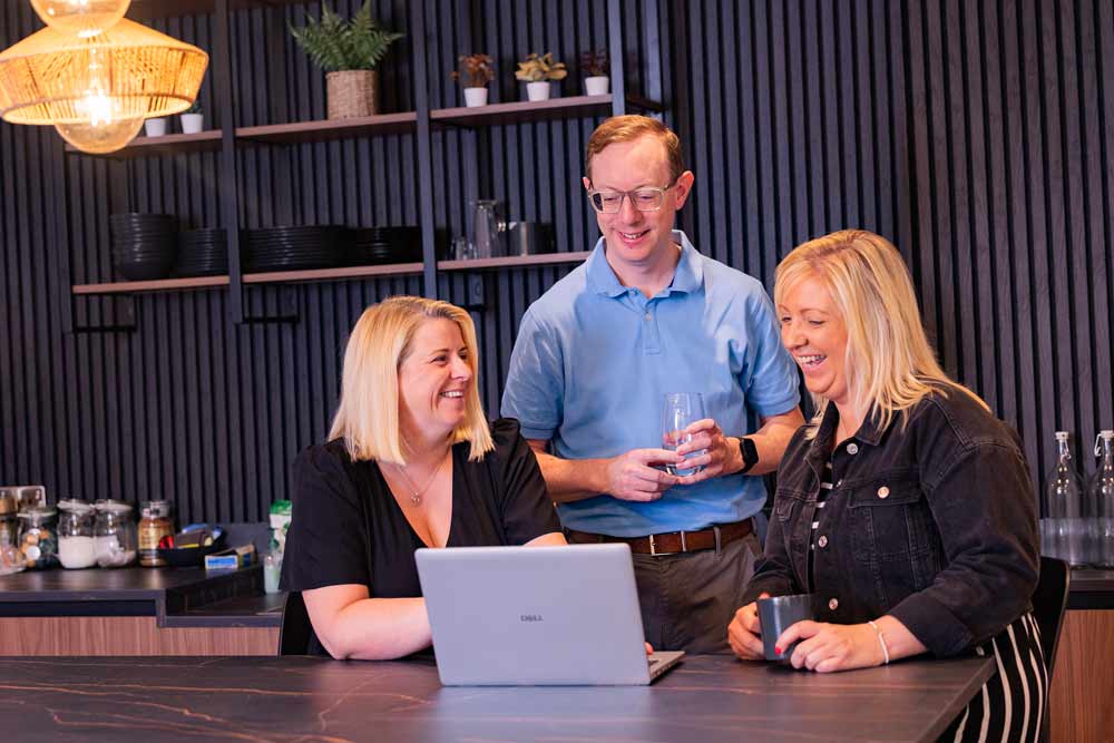 Three colleagues laughing over a laptop in the office kitchen